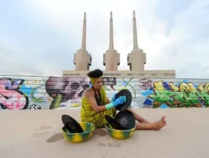 djatodocolor lavando vinilos al aire libre, rodeada de graffiti colorido y una fábrica industrial las tres chimeneas de fondo.