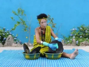 Retrato de la artista "djatodocolor" limpiando discos de vinilo, vestida de manera colorida, sentada al aire libre sobre una alfombra azul