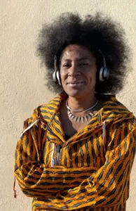 djatodocolor con peinado afro, auriculares y chaqueta estampada en tonos vibrantes, preparada para su actuación y destacando su identidad única.