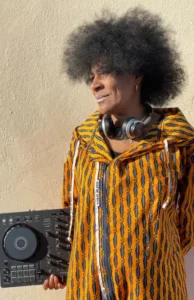 djatodocolor sostiene su mezcladora, vestida con chaqueta étnica vibrante y auriculares, mostrando su estilo y creatividad en la música.