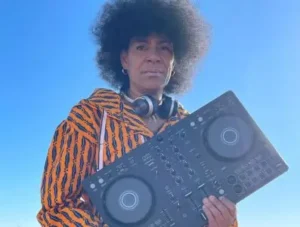 djatodocolor sostiene su mezcladora, vestida con chaqueta de estampado vibrante y auriculares, destacando bajo un cielo azul despejado.