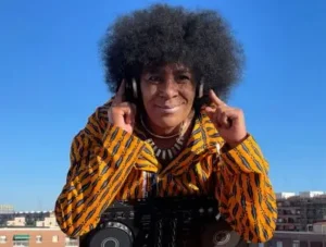 djatodocolor con auriculares y mezcladora, vestida con chaqueta vibrante, lista para crear en una escena al aire libre bajo un cielo azul.