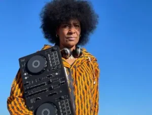 djatodocolor sostiene su mezcladora, con auriculares y chaqueta estampada vibrante, destacando bajo el cielo azul y preparada para su actuación.