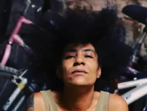 Primer plano de djatodocolor, DJ femenina con mirada introspectiva y cabello voluminoso, en un ambiente urbano con bicicletas al fondo.